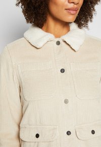 Chaqueta de pana beige con un suave collar de piel blanca. Cuenta con dos grandes bolsillos en el pecho y botones de metal en la parte delantera y en los bolsillos.