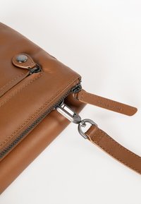Bruine leren tas met een ritssluiting, voorzien van een zilveren metalen trekker en een afneembare schouderband. Gladde textuur met zichtbare stiksels.