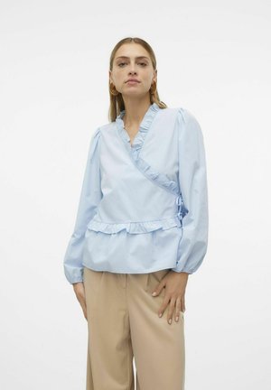 Lysblå indpakningsbluse med volangdetaljer, lange pufærmer og snoretræk. Sammen med beige bukser. Glat stoftekstur.