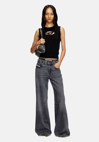 Haut sans manches noir avec des détails découpés, associé à un jean gris à pattes larges. Le mannequin tient un sac à main noir brillant, affichant un style urbain décontracté.
