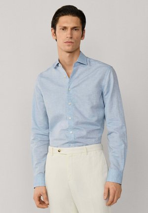 Man draagt een lichtblauw overhemd met lange mouwen, ingestopt in hooggetailleerde crèmekleurige broek, staand tegen een effen achtergrond.