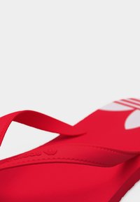 Φωτεινό κόκκινο σανδάλι Adidas με ανάγλυφο λογότυπο στο λουράκι και μεγάλο λευκό λογότυπο trefoil στην επιφάνεια του πέλματος, εμφανιζόμενο υπό γωνία σε απλό φόντο.