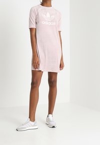 Krótka różowa sukienka z miękkiego materiału, z okrągłym dekoltem i krótkimi rękawami. Na przodzie znajduje się duże białe logo Adidas. Połączona z szarymi sneakersami.