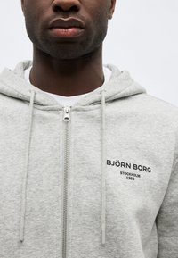 Κοντινό πλάνο ενός ατόμου που φοράει ανοιχτό γκρι ζακέτα Björn Borg με φερμουάρ, κορδόνια και μαύρο κείμενο στο στήθος.