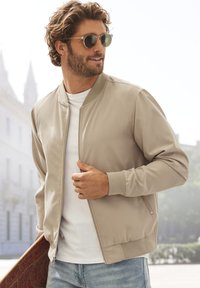 Beige Bomberjacke mit Reißverschluss und gerippten Bündchen, getragen über einem weißen T-Shirt. Das Model hält ein Skateboard und trägt runde Sonnenbrillen.