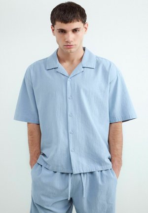 Mladý muž s krátkymi tmavými vlasmi, oblečený v svetlomodrú krátku košeľu na gombíky a zodpovedajúce nohavice so šnúrkou, stojaci s rukami v vreckách.