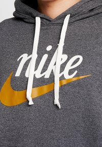 Grå hoodie med känguruficka, vita snören, med en stor vit "nike" logo och en orange swoosh-grafik. Mjuk textur.