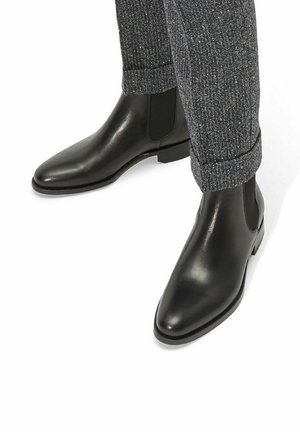 Botas negras de cuero hasta el tobillo con una punta afilada y paneles laterales elásticos, combinadas con pantalones grises texturizados arremangados en el tobillo.