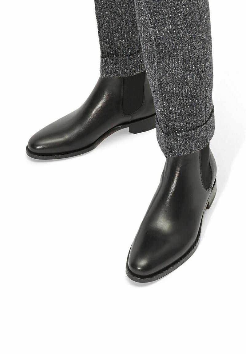 Botas negras de cuero hasta el tobillo con una punta afilada y paneles laterales elásticos, combinadas con pantalones grises texturizados arremangados en el tobillo.