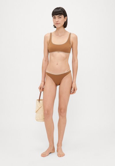 Smeđa bikini sa kontrastnim bijelim šavovima, s kvadratnim izrezom i donjim dijelom s visokim strukom. Model drži torbu od trske.