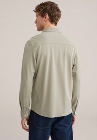 Hellgrünes langärmeliges Hemd aus strukturiertem Stoff, mit geknöpften Manschetten und spitzem Kragen, getragen zu dunkelblauen Jeans. Schlichtes Design, ohne Muster.