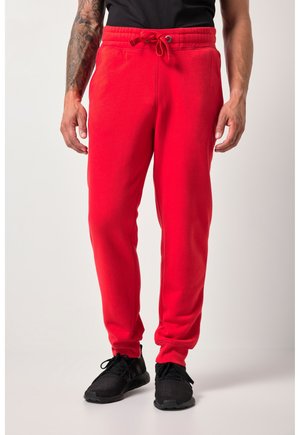 Mężczyzna ubrany w jaskrawoczerwone spodnie dresowe ze ściągaczem w talii i czarne buty sportowe, stojący na tle gładkiego, jasnego tła.