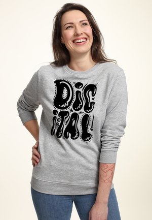Smilende kvinde med langt brunt hår iført grå sweatshirt med teksten "DIG IT ALL" i sorte boblebogstaver, står med hånden på hoften.