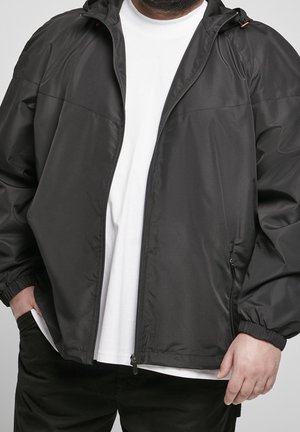 Adulto che indossa una giacca nera con zip sopra una camicia bianca, con le mani parzialmente nelle tasche, su uno sfondo semplice.