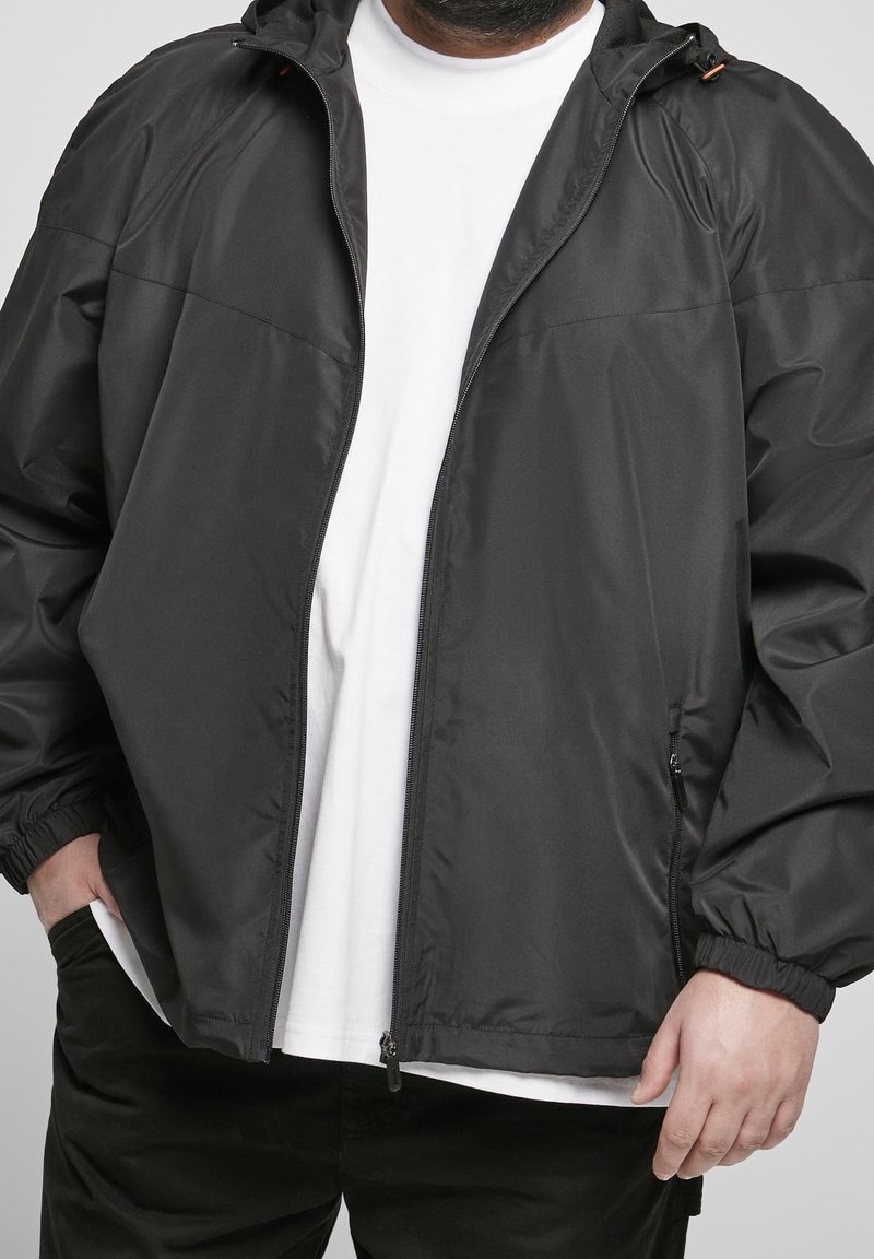 Adulte portant une veste noire zippée par-dessus une chemise blanche, les mains partiellement dans les poches, sur un fond uni.