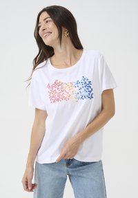 T-shirt en coton blanc arborant un design de dégradé pixelisé en rouge, rose, jaune et bleu. Manches courtes et encolure ronde.