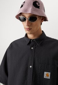 Camicia nera a maniche lunghe con texture, dotata di una tasca sul petto con il logo Carhartt. Abbinata a un cappello a secchiello lavorato a maglia rosa e occhiali da sole scuri.
