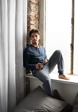 Chemise à boutonnière bleu marine à manches longues, pantalon de pyjama rayé gris et blanc, assis sur le rebord d'une fenêtre, tenant une tasse noire. Des coussins doux à proximité.