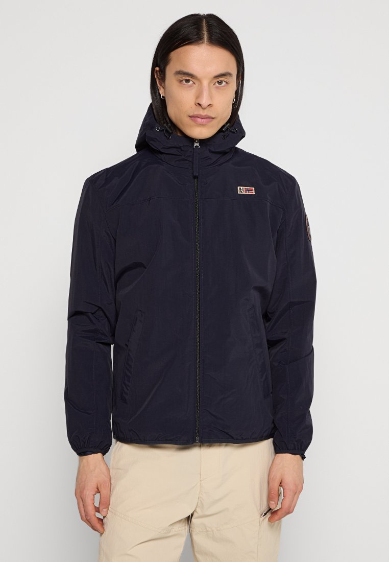 Uomo con capelli lunghi che indossa una giacca a zip con cappuccio blu navy e pantaloni beige, in piedi contro uno sfondo chiaro e uniforme.