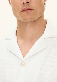 Camicia bianca a maniche corte con un tessuto testurizzato, colletto a punte e chiusura con un solo bottone. Un sottile dettaglio a motivo arricchisce il design.