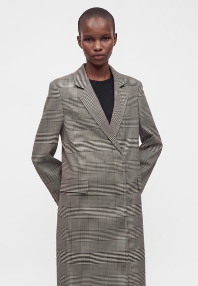 Personne aux cheveux courts portant un long blazer gris à carreaux par-dessus un haut noir, debout les mains derrière le dos sur un fond uni.