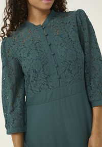 Abito in pizzo teal con maniche a tre quarti, corpetto in pizzo con motivi floreali e chiusura frontale con bottoni in tessuto. Parte inferiore liscia.