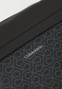 Borsetă din piele neagră, cu un model repetitiv al logo-ului în albastru închis, având un accent argintiu cu numele mărcii "Calvin Klein" pe față.