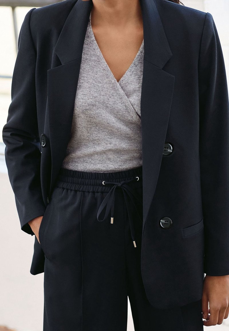 Blazer blu navy con bottoni grandi, abbinato a un top avvolgente grigio. I pantaloni neri con coulisse presentano una consistenza liscia e una vita elastica.