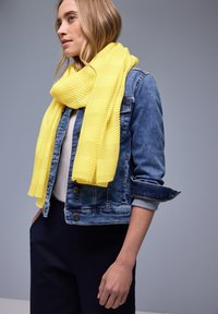 Femme portant une écharpe texturée jaune vif sur une veste en jean et un haut blanc, debout devant un fond gris clair uni.