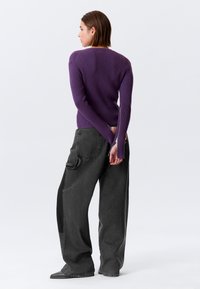 Pull violet côtelé à manches longues, associé à un pantalon gris foncé taille haute à jambes larges. Le mannequin est légèrement de profil.