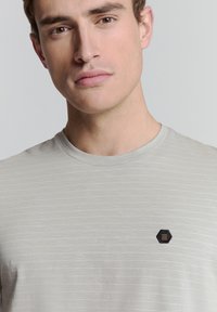 Lichtgrijze T-shirt met dunne horizontale strepen, ronde hals en een kleine zwarte hexagonale patch met drie horizontale lijnen op de borst.