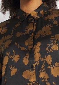 Camicia nera con motivo floreale dorato, colletto a punta e texture morbida. Presenta piccoli bottoni neri e una vestibilità rilassata.