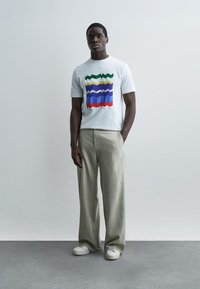 Mees, kes kannab valget t-särki värvilise siksakmustriga, beeže laiapõlvalisi pükse ja valgeid tossusid, seisab ühevärvilise heleda tausta ees.