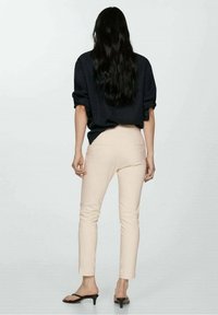 Chemise noire oversize, pantalon beige ajusté et sandales à talons noirs vues de dos. Cheveux longs et ondulés, laissés libres sur les épaules.