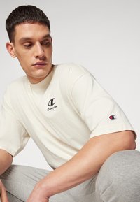 Cremfarbendes T-Shirt mit kurzen Ärmeln, schwarzem Logo auf der Brust und gesticktem Patch am Ärmel. Weicher Stoff, lockere Passform, minimalistisches Design.