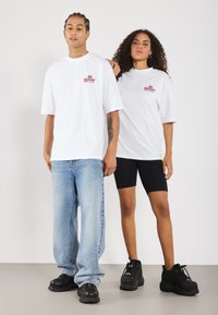Hvide oversized t-shirts med logo accenter, parret med blå baggy jeans og sorte shorts, begge iført sorte chunky sneakers.