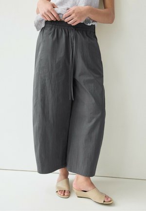 Personne portant un pantalon large raccourci gris foncé avec taille élastique et cordon, debout contre un mur clair, sandales compensées beige à enfiler.
