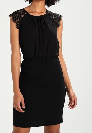 Femme portant une robe noire sans manches avec des manches en dentelle et une taille froncée, debout sur un fond clair uni.
