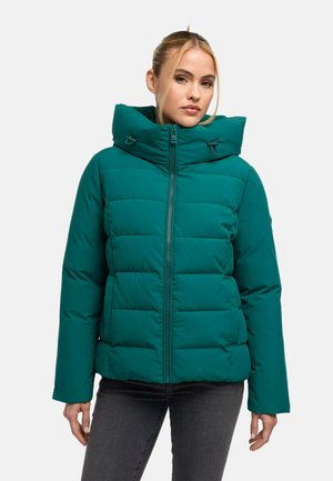Frau trägt eine türkisfarbene gesteppte Jacke mit hohem Kragen und Reißverschluss, steht vor einem einfarbigen hellen Hintergrund.