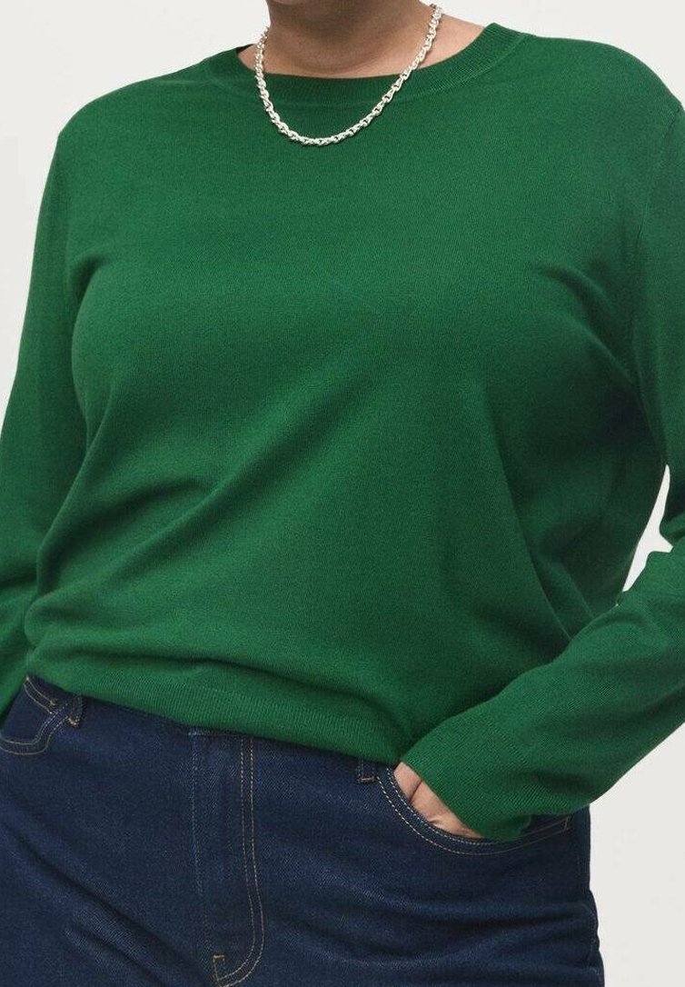 Personne portant un pull vert à manches longues, jean bleu foncé, collier chaîne argentée, une main dans la poche, sur un fond uni.