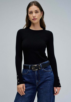 Top negro de manga larga ajustado con cuello redondo y detalles de botones en los puños, combinado con jeans de mezclilla azul de cintura alta y un cinturón negro.