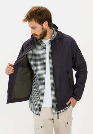 Marineblaues leichter Jacke mit Kapuze, Reißverschluss vorne und Mesh-Futter. Darüber trägt er ein grün-weiß kariertes Hemd, kombiniert mit beigen Hosen.