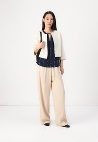 Gräddfärgad kort jacka över en marinblå blus med prickar, kombinerad med beige vida byxor och svarta spetsiga ballerinas. Modellen håller en svart väska.