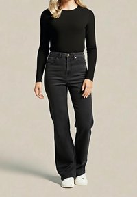 Femme portant un haut noir à manches longues, un jean noir taille haute et des baskets blanches, debout devant un fond beige uni.
