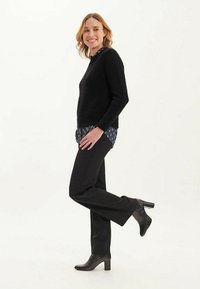 Pull en tricot noir avec un col rond, associé à un pantalon sombre et des bottes à talons noires. Un chemisier à motifs est visible en dessous.