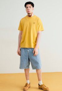 Tricou galben din bumbac, șorturi din denim albastru, încălțări maro, șosete albe. Design simplu, confortabil, cu un mic logo pe piept.