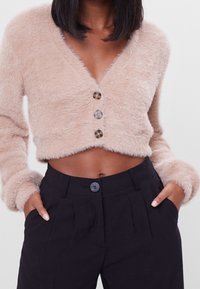 Ljusbeige kortärmad cardigan med fluffig textur, V-urringning och tre runda knappar. Matchad med högmidjade svarta byxor.