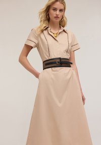 Beige cotton dress with a collared neckline, button front, and decorative stitching. Worn with a black woven belt and chunky gold necklace.