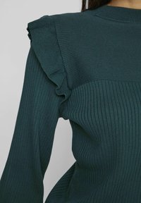Pull en maille côtelée vert foncé avec détail à volants sur l'épaule, ajusté au niveau du torse, porté sur le haut du corps sur un fond uni.
