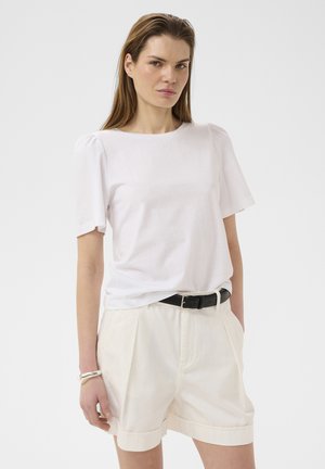 Frau trägt ein schlichtes weißes Kurzarmshirt, cremefarbene Shorts mit hohem Bund und Umschlag, schwarzen Gürtel und silbernes Armband, steht vor weißem Hintergrund.